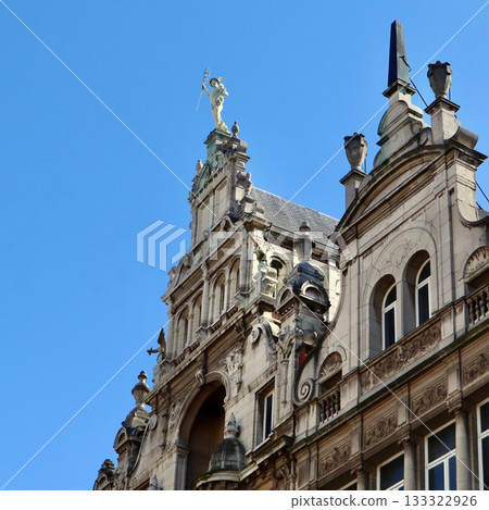 Stone building adorned with intricate carvings is topped with a statue of Mercury, the Roman god of commerce, overlooking a clock below against a clear blue sky. Antwerp, Belgium Stone building adorned with intricate carvings is topped with a statue of Mercury, the Roman god of commerce, overlooking a clock below against a clear blue sky. Antwerp, Belgium 133322926