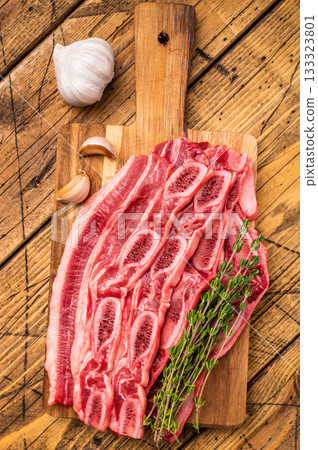 Overhead of uncooked cross-cut short ribs on a cutting board with herbs, set against a dark wood surface. Overhead of uncooked cross-cut short ribs on a cutting board with herbs, set against a dark wood surface. 133323801