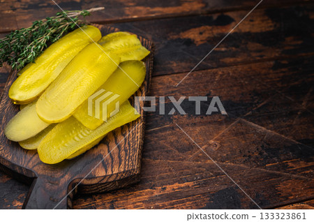 Yellow pickled cucumber slices on rustic wooden cutting board with thyme and peppercorns, top view of brined savory snack. 133323861