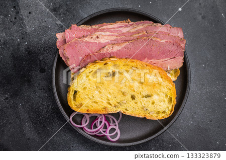 Smoked pastrami brisket slices in a hearty sandwich with seed crusted bread, onions and pickles on dark stone surface. 133323879