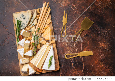Rustic cheese platter with nuts herbs and breadsticks displayed on a wooden board set against a rich dark textured backdrop. 133323888