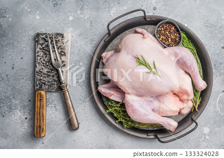 Raw whole chicken on a rustic metal tray with fresh rosemary, thyme, and peppercorns on a gray concrete background, ready for cooking. 133324028