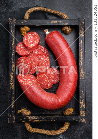 Sliced and whole Turkish sucuk (sujuk) in a rustic tray with a fork. Traditional spicy dry sausage on a dark background. 133324053