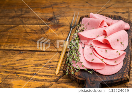 Slices of pink bologna lunch meat served on a rustic dark wooden cutting board. Dark, moody background with copy space. 133324059