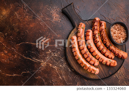Grilled and cut sausages (frankfurters/wieners) seasoned with thyme, served on a dark wooden board with Himalayan salt. Ready meal. 133324060