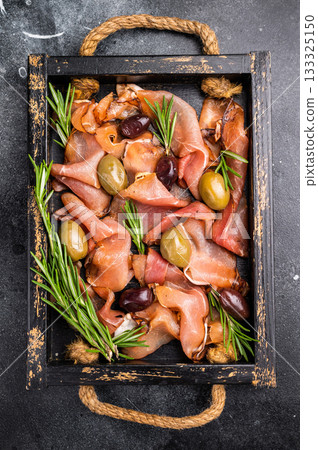 Prosciutto ham or Jamon Serrano with rosemary and olives in wooden tray. black background. top view Prosciutto ham or Jamon Serrano with rosemary and olives in wooden tray. black background. top view 133325150