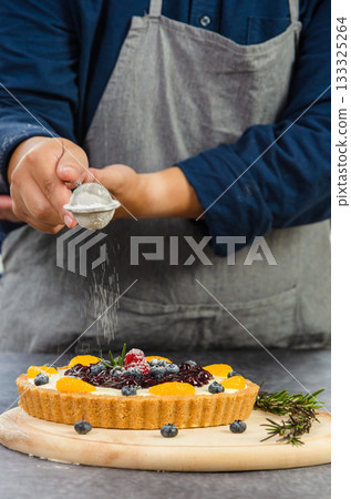 Baker in navy shirt and gray apron sifting icing sugar through fine mesh sieve onto blueberries cheese pie on wooden board in kitchen 133325264