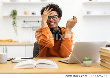 A young man wearing glasses looks stressed while holding a game controller and sitting at a desk with a laptop and books. 133325733