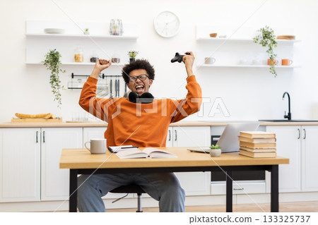 A young man celebrates a win while gaming in his kitchen. He is holding a game controller and is very excited. A young man celebrates a win while gaming in his kitchen. He is holding a game controller and is very excited. 133325737