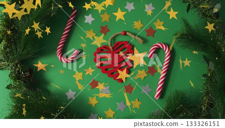 Red wooden heart ornament lying on green table, with candy canes, pine garland, star confetti 133326151