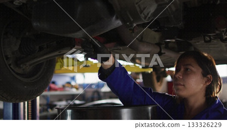 Female mechanic wearing blue coverall reaching under raised chassis in workshop, with oil drain pan Female mechanic wearing blue coverall reaching under raised chassis in workshop, with oil drain pan 133326229