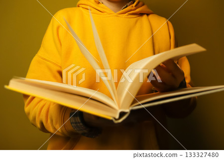 Person in yellow hoodie reading open book against warm brown background. a woman reading at home. A person's hand is holding an open book with slightly yellowed paper pages 133327480