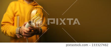 Person in yellow hoodie holding large glowing vintage-style LED light bulb against warm background. Hand holding a filament light bulb isolation on dark yellow background. Innovation concept 133327482