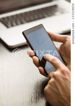 Hands are holding black smartphone and tapping screen above laptop on wooden desk in flat design 133329059