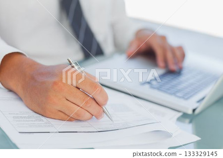 Businessman writing on documents while using laptop 133329145