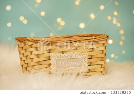A natural wicker basket with a lace-edged Home label stands on a fluffy white surface against a soft blue background with warm bokeh lights. comfort, coziness, and home decor aesthetics 133329250