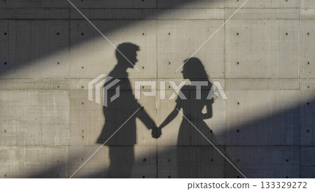 Silhouettes of a man and woman holding hands on a textured concrete wall in streaks of bright sunlight. 133329272