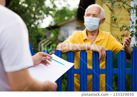 Grandfather in protective mask talking to census agent standing at fence of his country house 133329830