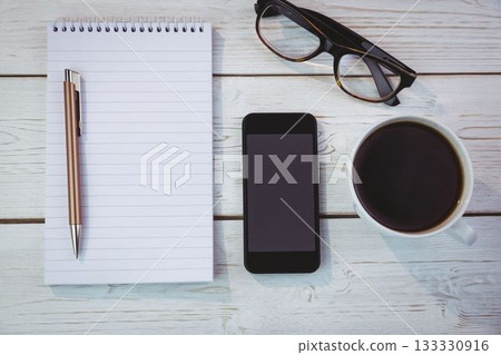 Flat lay is showing spiral notebook with pen on white table with smartphone, eyeglasses, coffee mug 133330916
