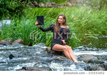 female sitting on a stone in the river and making a video call on a digital tablet female sitting on a stone in the river and making a video call on a digital tablet 133331852