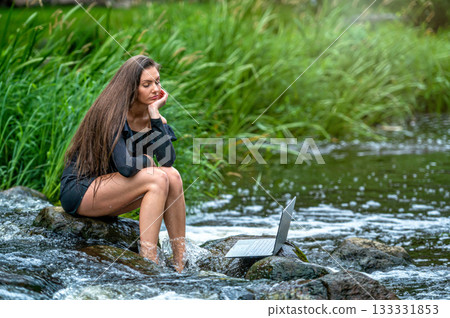 woman freelancer sitting on a rock in the river and using a laptop, travel or vacation work concept 133331853
