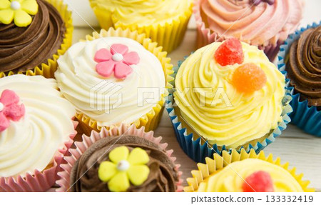 Flat design showing cupcakes on wood surface with liners, buttercream, fondant flowers, gumdrops Flat design showing cupcakes on wood surface with liners, buttercream, fondant flowers, gumdrops 133332419