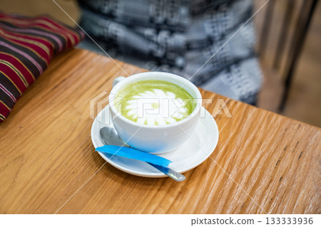 A warm cup of matcha latte rests on a white saucer atop a wooden table, with white foam forming a simple floral design in the center. A warm cup of matcha latte rests on a white saucer atop a wooden table, with white foam forming a simple floral design in the center. 133333936