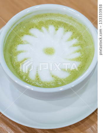 A warm cup of matcha latte rests on a white saucer atop a wooden table, with white foam forming a simple floral design in the center. A warm cup of matcha latte rests on a white saucer atop a wooden table, with white foam forming a simple floral design in the center. 133333938