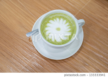 A warm cup of matcha latte rests on a white saucer atop a wooden table, with white foam forming a simple floral design in the center. A warm cup of matcha latte rests on a white saucer atop a wooden table, with white foam forming a simple floral design in the center. 133333941