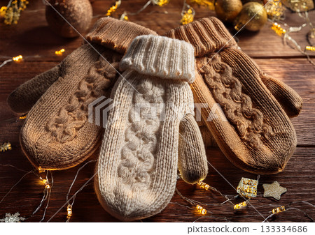 Brown hand-knitted mittens on a wooden table with Christmas ornaments and illumination lights. Winter gift. 133334686