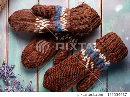 A pair of hand-knitted brown mittens against a background of pale blue aged wood grain and star-shaped Christmas ornaments 133334687
