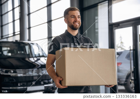 With a box, smiling. Delivery man or loader is near the car With a box, smiling. Delivery man or loader is near the car 133336163