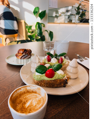 Avocado toast with mozzarella, cherry tomatoes and basil served on plate next to cappuccino, cinnamon bun and glass of water on wooden table in cafe with customer sitting in background. Cafe interior Avocado toast with mozzarella, cherry tomatoes and basil served on plate next to cappuccino, cinnamon bun and glass of water on wooden table in cafe with customer sitting in background. Cafe interior 133336204