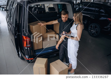 Woman is helping delivery man with products, holding notepad, loader is working with boxes from automobile Woman is helping delivery man with products, holding notepad, loader is working with boxes from automobile 133336287