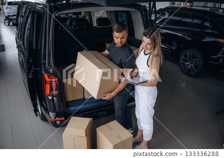 Woman is helping delivery man with products, holding notepad, loader is working with boxes from automobile Woman is helping delivery man with products, holding notepad, loader is working with boxes from automobile 133336288