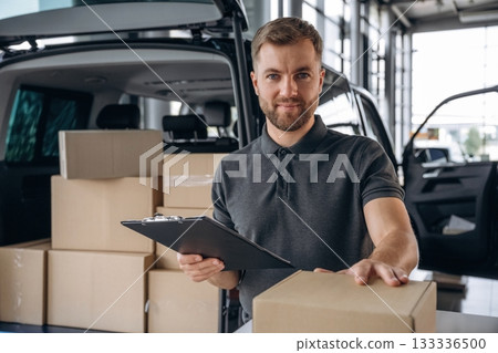 Positive facial expression, holding notepad. Delivery man or loader is near the car with a box Positive facial expression, holding notepad. Delivery man or loader is near the car with a box 133336500