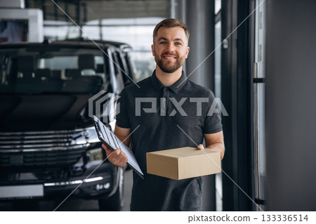 Standing, holding notepad. Delivery man or loader is near the car with a box 133336514