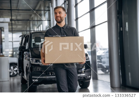 With a box, smiling. Delivery man or loader is near the car With a box, smiling. Delivery man or loader is near the car 133336527