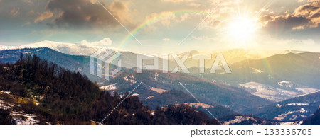 panorama of rolling hills and mountain ridge with snow cowered tops at sunset. beautiful landscape in springtime cold weather in evening light. timeless nostalgia book cover with rainbow 133336705