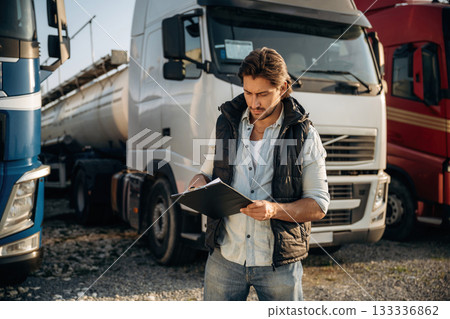 Black notepad, standing and holding it. Truck driver is near the transport outdoors Black notepad, standing and holding it. Truck driver is near the transport outdoors 133336862
