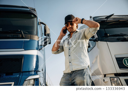 In the casual clothes and in black cap. Truck driver is near the transport outdoors In the casual clothes and in black cap. Truck driver is near the transport outdoors 133336943