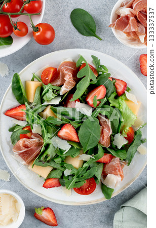 Italian arugula salad with prosciutto, strawberries, melon, cheese on gray background. Top view.  133337537