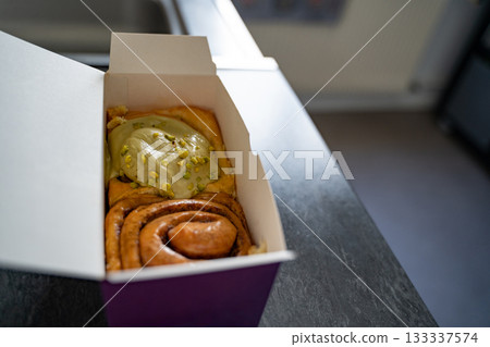 Classic and pistachio cinnamon rolls arranged in a takeout cardboard box, viewed from above. Concept of bakery, pastry, takeaway, breakfast, indulgence. Classic and pistachio cinnamon rolls arranged in a takeout cardboard box, viewed from above. Concept of bakery, pastry, takeaway, breakfast, indulgence. 133337574