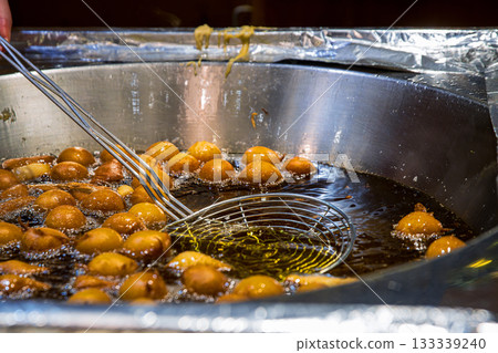Street food vendor stirring and frying golden dough balls stirring them with metal skimmer tool, in sizzling hot oil at a winter market stall. Street food vendor stirring and frying golden dough balls stirring them with metal skimmer tool, in sizzling hot oil at a winter market stall. 133339240