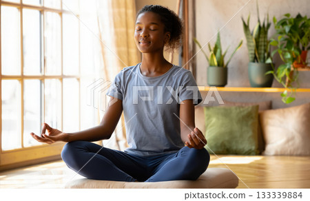 Mental health and self-care concept with a young woman sits in lotus position on a yoga mat at home, meditating with her eyes closed and holds her hands in Mudra. Mindfulness, inner calm and wellness 133339884