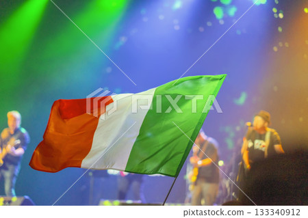 Irish tricolor flag waving at night concert during St. Patrick Day celebration, symbolizing pride and heritage. Concept of national unity, culture, festive identity 133340912