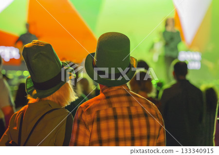 Couple in green hats enjoying St. Patrick Day celebration at club with Irish flag lights on stage. Concept of unity, tradition, national pride 133340915
