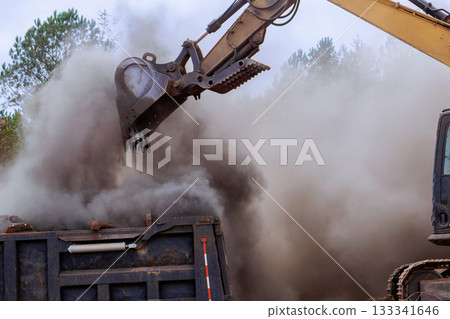 Excavator is lifting dirt debris from ground into dump truck, creating dusty cloud 133341646
