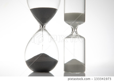 A sleek hourglass with dark sand flowing against a stark white background A sleek hourglass with dark sand flowing against a stark white background 133341973