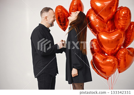 Man posing for a photo while make a proposal for his girlfriend. Studio full length shot of man holding an engagement ring while standing near bunch of heart-shaped red ballons. Brunette woman and Man posing for a photo while make a proposal for his girlfriend. Studio full length shot of man holding an engagement ring while standing near bunch of heart-shaped red ballons. Brunette woman and 133342331